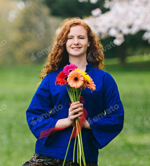 Preview: Portrait of young woman with long wavy red hair holding bunch of flowers in park