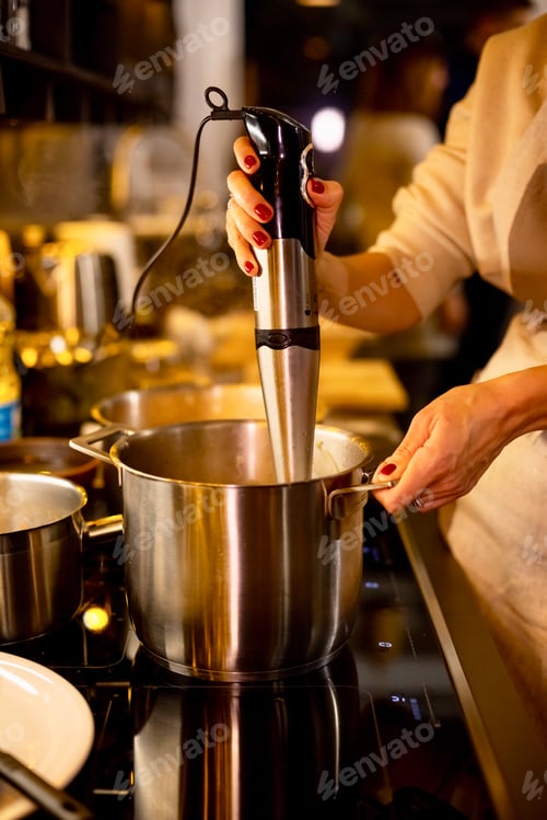 Preview: Woman in linen apron cooking with blender pumpkin soup in saucepan on induction oven in dark kitchen
