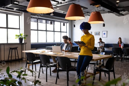 Preview: Biracial female creative sitting on desk using tablet in casual office