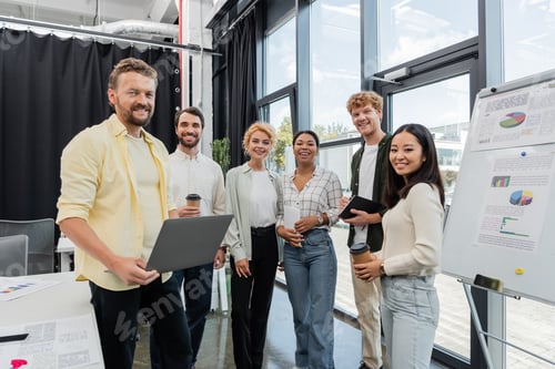 Preview: multiethnic business team with paper cups and gadgets smiling at camera near flip chart