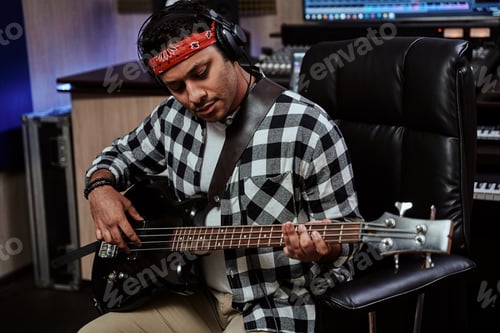 Preview: Portrait of focused young man, male artist in headphones playing guitar while sitting in recording