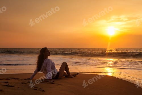 Preview: Silhouette of slim woman sitting on tropical beach at ocean sunset, rear side view