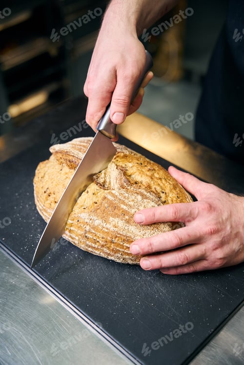 Preview: Cropped male chef cutting bread with knife on chopping board at table on kitchen