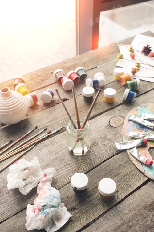 Preview: Paint brushes in a water and scattered containers with poster paints