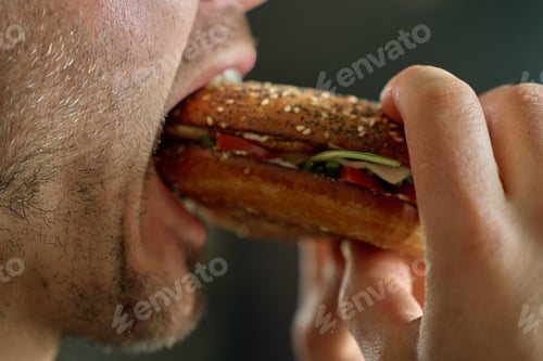 Preview: Mouth taking bite from crunchy bagel sandwich with greens and tomato