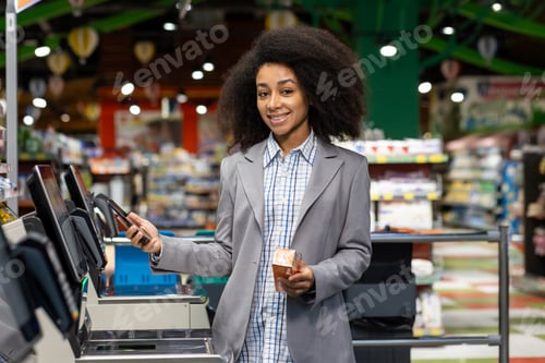 Preview: A young woman with curly hair and a gray blazer smiles while using a selfcheckout in a grocery