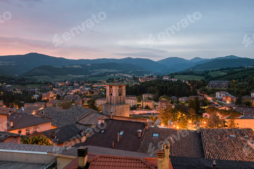 Preview: High angle shot of a lot of buildings with lights surrounded by green trees in Cascia, Italy