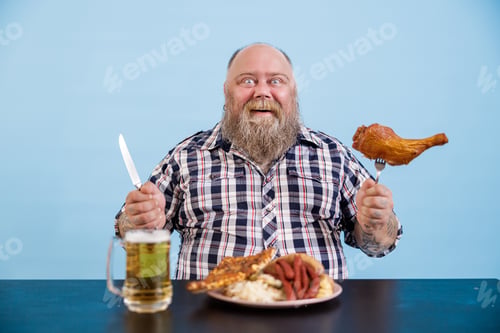 Preview: Smiling man with overweight holds chicken leg sitting at table on light blue background