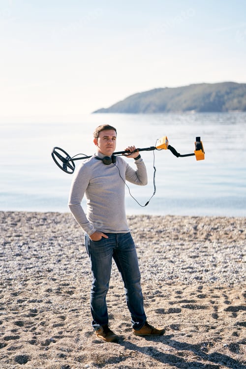 Preview: Man with headphones around his neck stands on the seashore with a metal detector on his shoulder