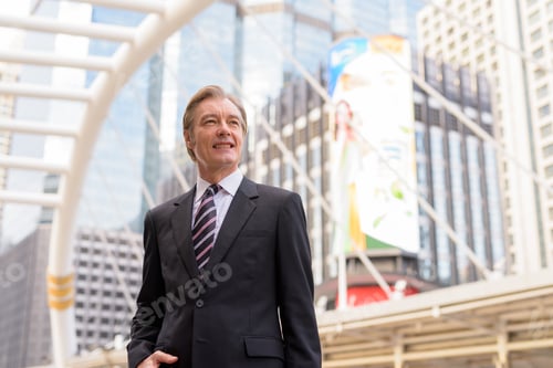Preview: Happy mature handsome businessman in suit thinking at skywalk bridge in the city