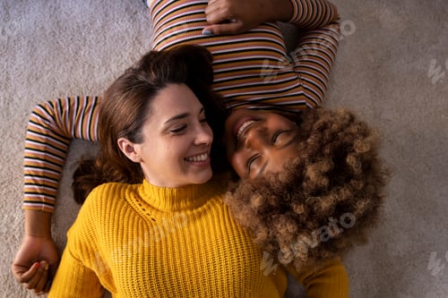 Preview: Top view of lovely multiracial lesbian couple lying on a carpet looking aat each other. Two