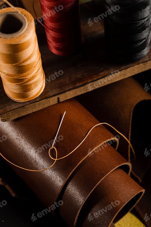 Preview: Close-up thread and needle on brown leather roll inside artisan workshop, handmade leather production