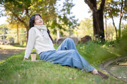 Preview: Asian woman in scarf closing eyes leaning back sitting leisurely with coffee on grass in garden park
