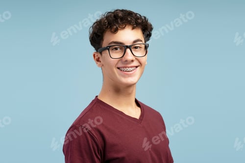 Preview: Smiling, happy teenager boy having braces wearing stylish eyeglasses isolated on blue background
