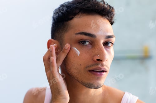 Preview: Biracial transgender man looking in mirror and applying face cream in bathroom