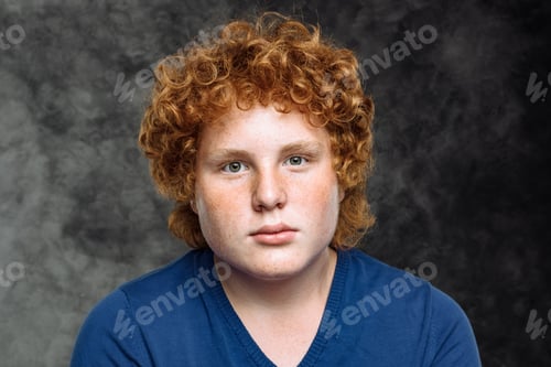 Preview: Studio portrait of deep curly red-haired freckled teenager in blue shirt on gray background