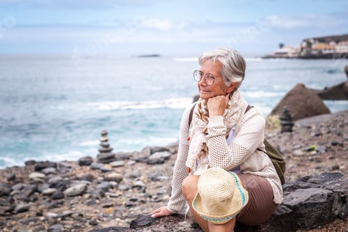 Preview: Attractive relaxed senior woman sitting on a pebble beach looking away pensive