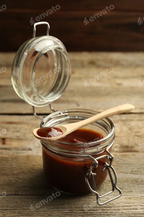 Vista previa: Sabrosa salsa barbacoa en tarro y cuchara sobre mesa de madera