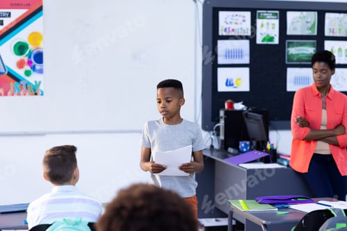 Preview: Diverse female teacher and pupils sitting at desks listening to schoolboy read to class