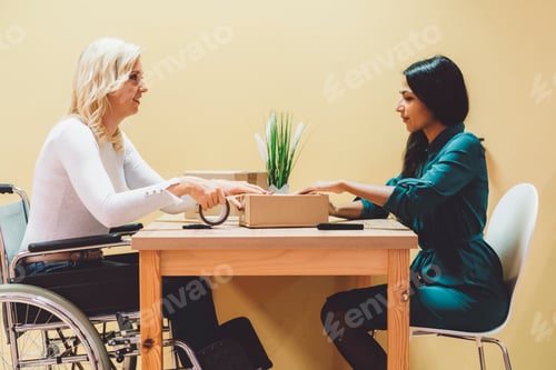 Preview: Indian woman and caucasian woman on wheelchair packaging a parcel