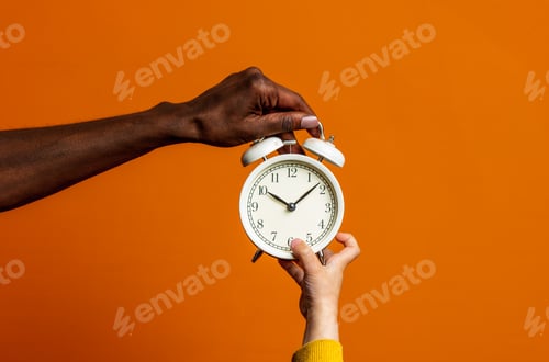 Preview: Black female hand and white child are holds alarm clock on orange background