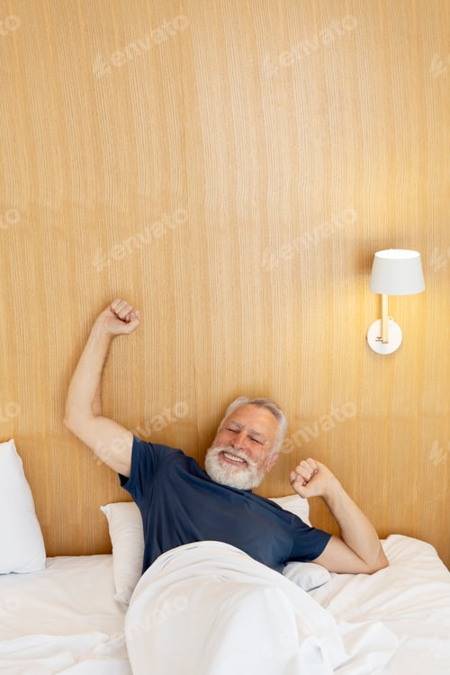 Preview: Positive elderly gray haired man with beard lying in bed, awake, stretching in hotel, inside