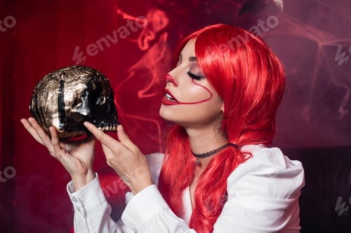 Preview: woman with red hair and clown makeup holding golden skull near spiderweb on dark red background
