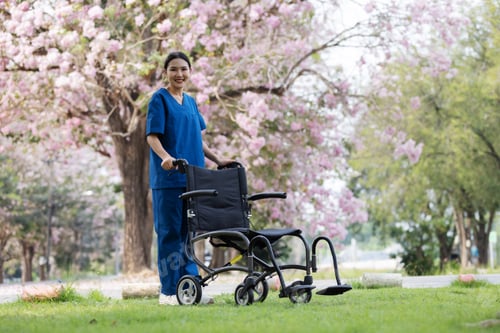 Vorschau: Eine fröhliche Krankenschwester in blauem Peeling steht hinter einem leeren Rollstuhl in einem ruhigen Park