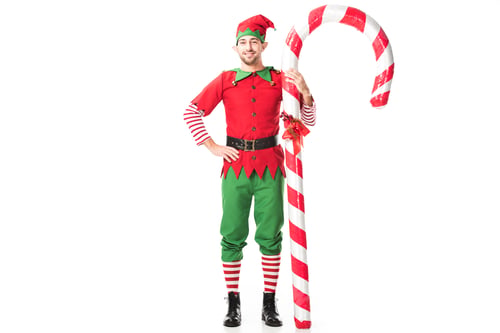 Preview: smiling man in christmas elf costume with hand on hips standing near big candy cane isolated on