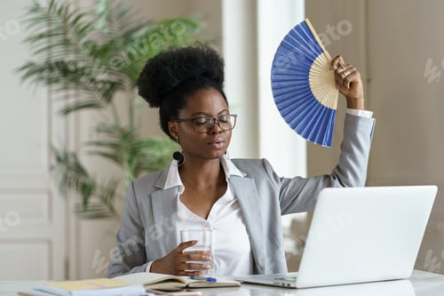 Preview: Exhausted businesswoman tired of heat waving paper fan for fresh air at workplace work on laptop