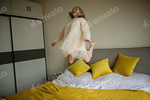 Preview: Beautiful little girl in stylish dress jumping on the bed with white blanket at home. Stay at home