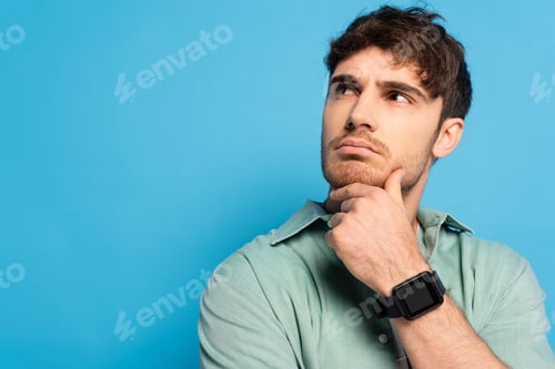 Preview: thoughtful young man looking away while touching chin on blue