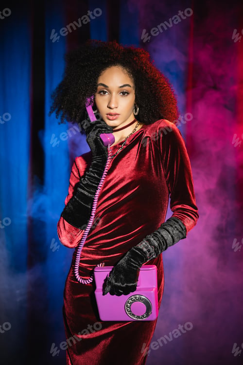 Visualização: Trendy african american woman in dress talking on telephone on red background with blue light and