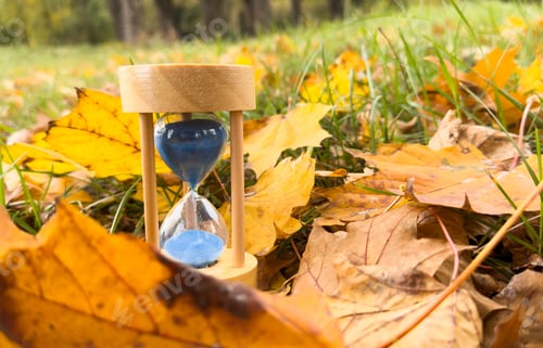 Preview: A blue sand clock is sitting on the ground in a field of fallen brown leaf. Concept of fall season