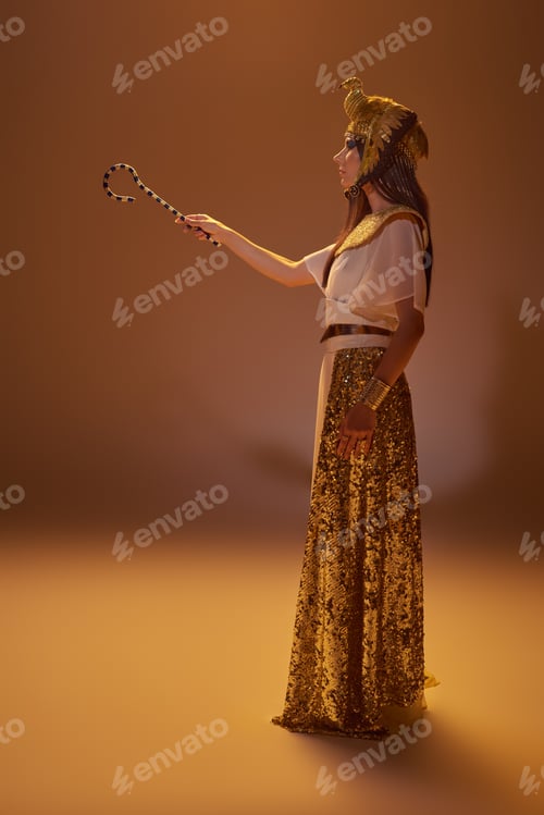 Preview: Side view of elegant woman in egyptian attire holding crook while standing on brown background