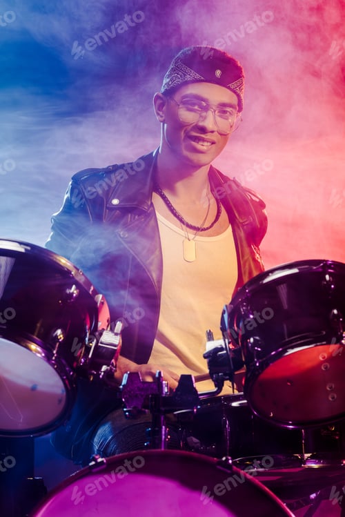Preview: cheerful mixed race male musician sitting behind drum set on stage with smoke and dramatic lighting