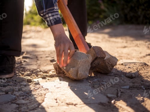 Preview: close up hand in shirt holding a hammer and crashing the stone against sun outdoor