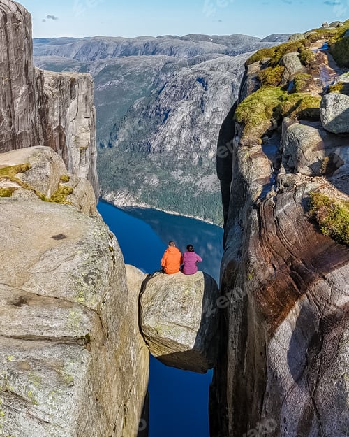 Preview: Two hikers sit on a rock formation that bridges the gap between two towering cliffs in Norway