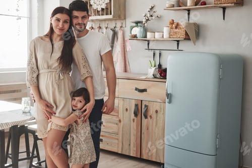 Preview: Cute family photo of pregnant mother, father and their daughter. Standing at the kitchen
