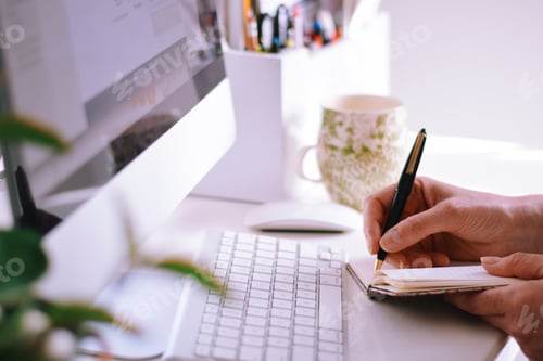 Preview: Woman sitting at a desk in modern home office in front of a computer and writing notes in notebook