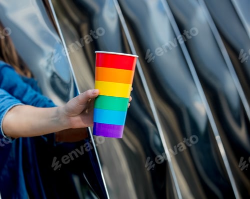 Preview: hand holding a cup in the color of the LGBT flag on the background of metal construction