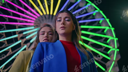 Preview: Fashion models looking camera at neon illuminated ferris wheel. Two girls posing