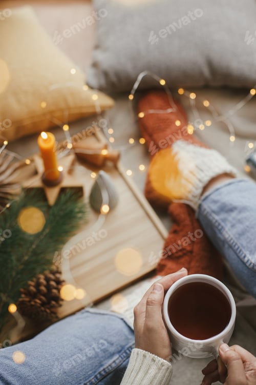 Preview: Woman hands with warm cup of tea with golden lights, christmas stars, pine, candle.Winter hygge