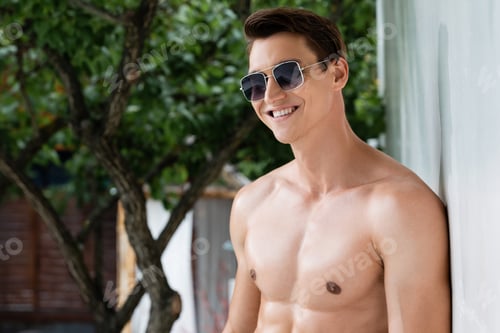 Preview: positive shirtless man in sunglasses smiling near wall outdoors