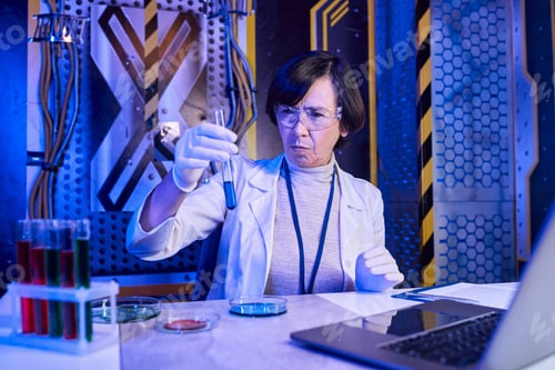 Preview: woman scientist in goggles looking at test tube with liquid near laptop in futuristic laboratory