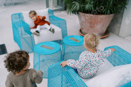 Preview: Little girls sit on blue wicker chairs near a large green bush in a clay pot. Back view