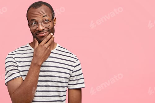 Preview: Horizontal shot of attractive dark skinned male with satisfied expression holds hand above chin, loo