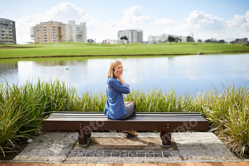 Preview: Rear view shot of a blonde woman talking on her phone on the park bench