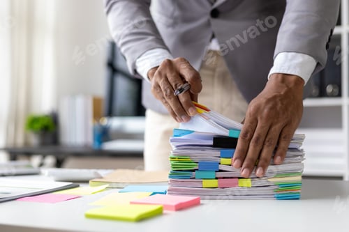 Preview: Man searching for documents in stack of papers on desk in office.