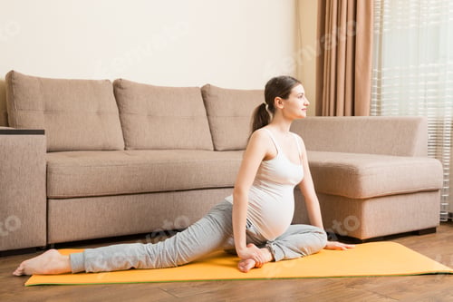 Preview: beautiful young pregnant woman sitting near sofa in yoga pose at the home